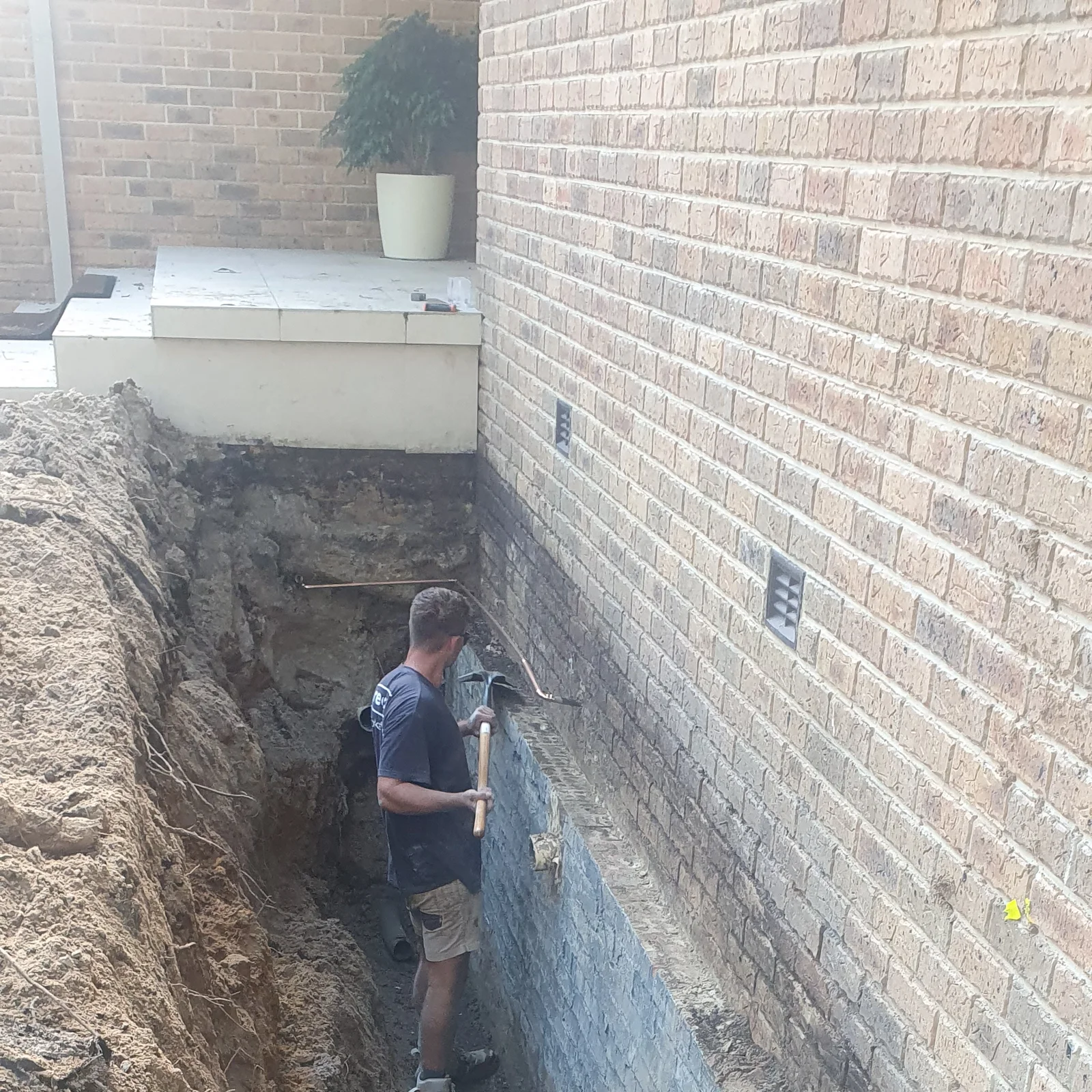 Hand-digging a deep drainage trench alongside a brick house foundation